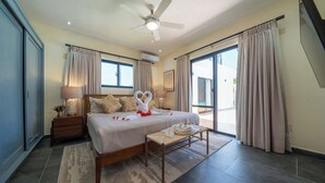 Habitación de hotel con cama, ventilador de techo, mesita de noche, banca y vistas al exterior.
