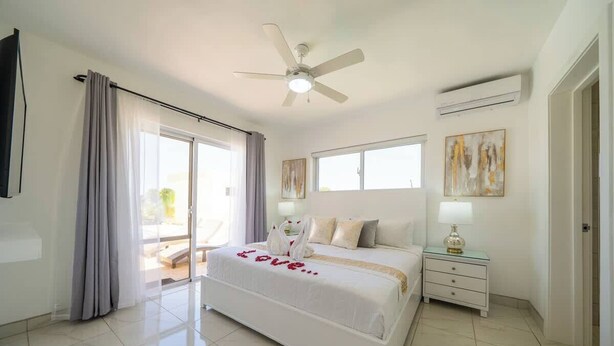 Un dormitorio con cama, ventilador de techo, mesita de noche, lámpara y un cuadro en la pared.