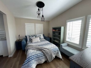 Quarto com cama, um banquinho, estante de livros e janela com persianas.