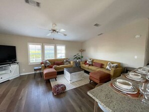 Uma sala de estar ampla com ventilador de teto, dois sofás, uma mesa de centro e um espaço de jantar com pratos e copos.
