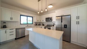 A modern kitchen with white cabinets, a central island, stainless steel appliances, and pendant lighting. - Location • Vibe • Style (Sosúa)