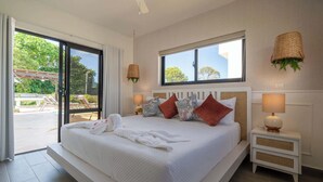 A large bed with white linens, a window with a view, a ceiling fan, and a small bedside table with a lamp. - Modern Villa · Pool, Jacuzzi & Pool Table · Sosúa (Sosúa)