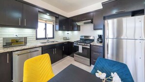 A modern kitchen with dark cabinets, stainless steel appliances, and a yellow and blue chair. - Modern 2BR Villa · Private Pool · Casa Linda (Sosúa)