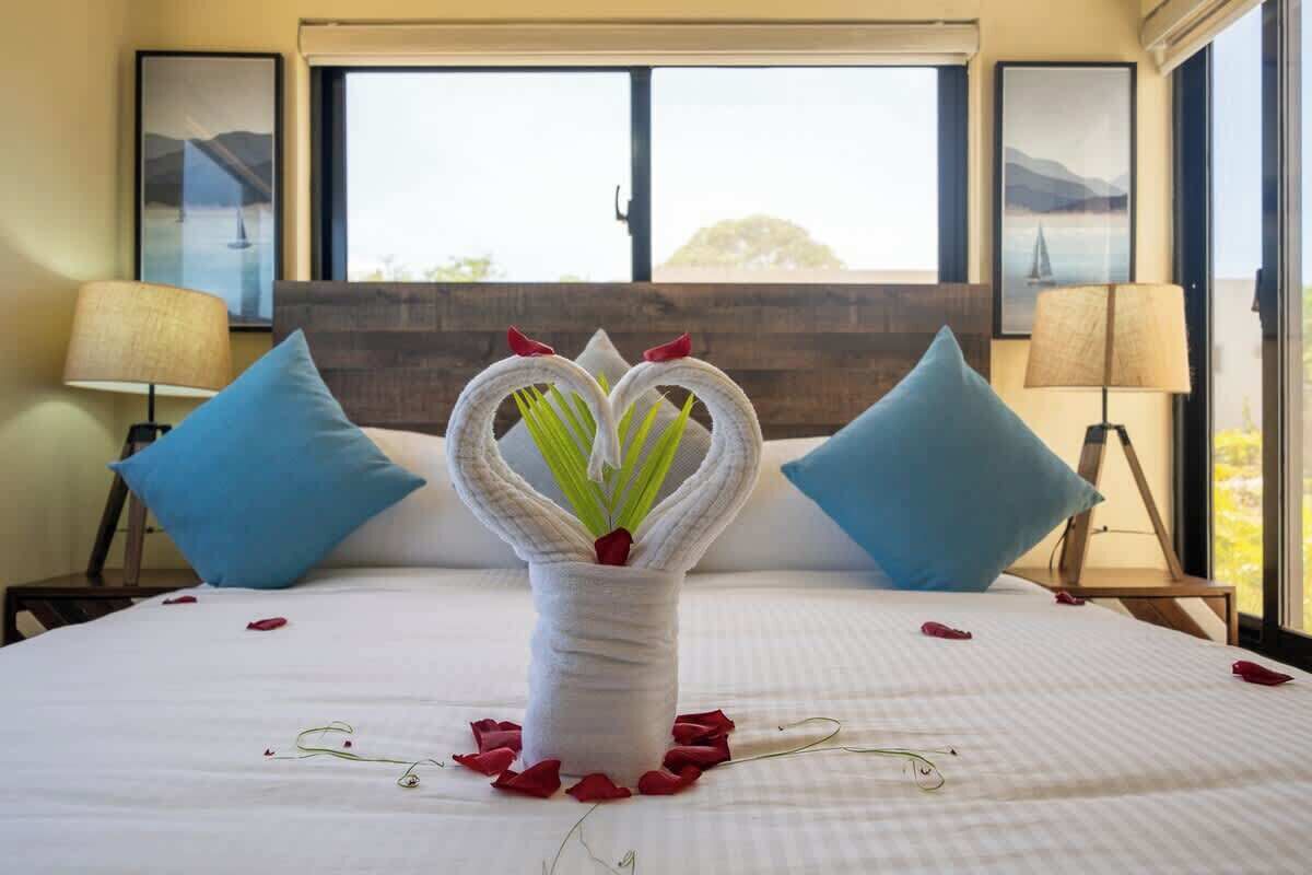 A bed with a white bedspread, two blue pillows, a vase with a floral arrangement, and a window with a view of a bridge and mountains.