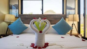 A bed with a white bedspread, two blue pillows, a vase with a floral arrangement, and a window with a view of a bridge and mountains.