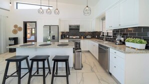 Eine moderne Küche mit einer zentralen Kochinsel, Barhocker, hängenden Pendelleuchten und weißen Schränken.