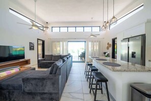 Una sala moderna con un televisor de pantalla plana grande, un sofá, un comedor con taburetes de bar y una cocina con refrigerador.