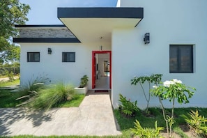 Ein modernes Haus mit weißer Fassade, einer roten Tür und einem kleinen Garten mit verschiedenen Pflanzen.