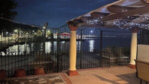 Ein Terrassenbereich im Freien bei Nacht mit Blick auf ein Gewässer, einem Zaun und einer Pergola mit Lichterketten.