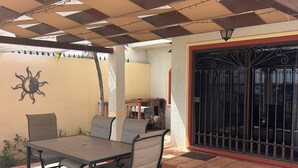 Eine Außenpatio mit Tisch und Stühlen, ein überdachter Bereich mit gemustertem Dach und ein eingezäunter Eingang.