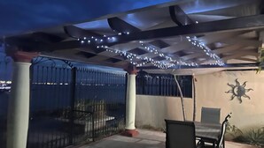 Ein überdachter Patio-Bereich mit Tisch und Stühlen, Lichterketten, einer dekorativen Sonne und Blick auf eine Stadtlandschaft.