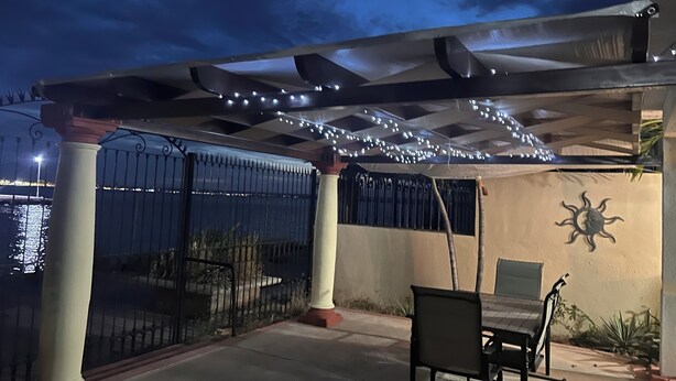 Eine überdachte Terrasse mit einer Pergola, Lichterketten, einem Tisch und Stühlen, einer Zierwand und Blick auf das Wasser.
