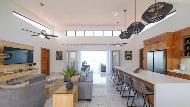 A modern living room with a sofa, coffee table, and TV, a kitchen with a fridge and bar seating, and a large window.
