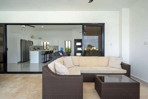 Ein modernes Wohnzimmer mit einer Eckcouch, einem Glastisch und einer Schiebetür zu einer Küche und einem Essbereich.