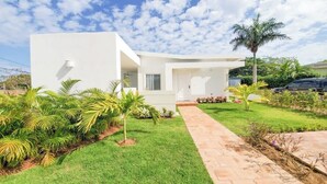 Uma casa moderna e branca com calçada pavimentada, rodeada por vegetação exuberante e palmeiras.