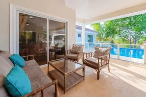 A balcony with wicker furniture, a glass table, and a view of a swimming pool and greenery. - Ocean View Luxe Retreat • 4BR • Pool & Serenity (Sosúa)