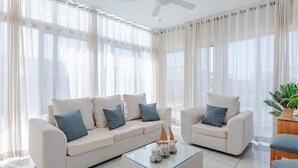 Ein modernes Wohnzimmer mit einer weißen Sofagarnitur, einem Glastisch und transparenten Vorhängen.