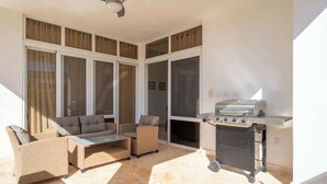 Eine sonnendurchflutete Terrasse mit Korbmöbeln, einem Grill und großen Fenstern.