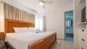 Ein Hotelzimmer mit einem hölzernen Bett, einem Deckenventilator, einem Fernseher und einem durch eine offene Tür einsehbaren Badezimmer.