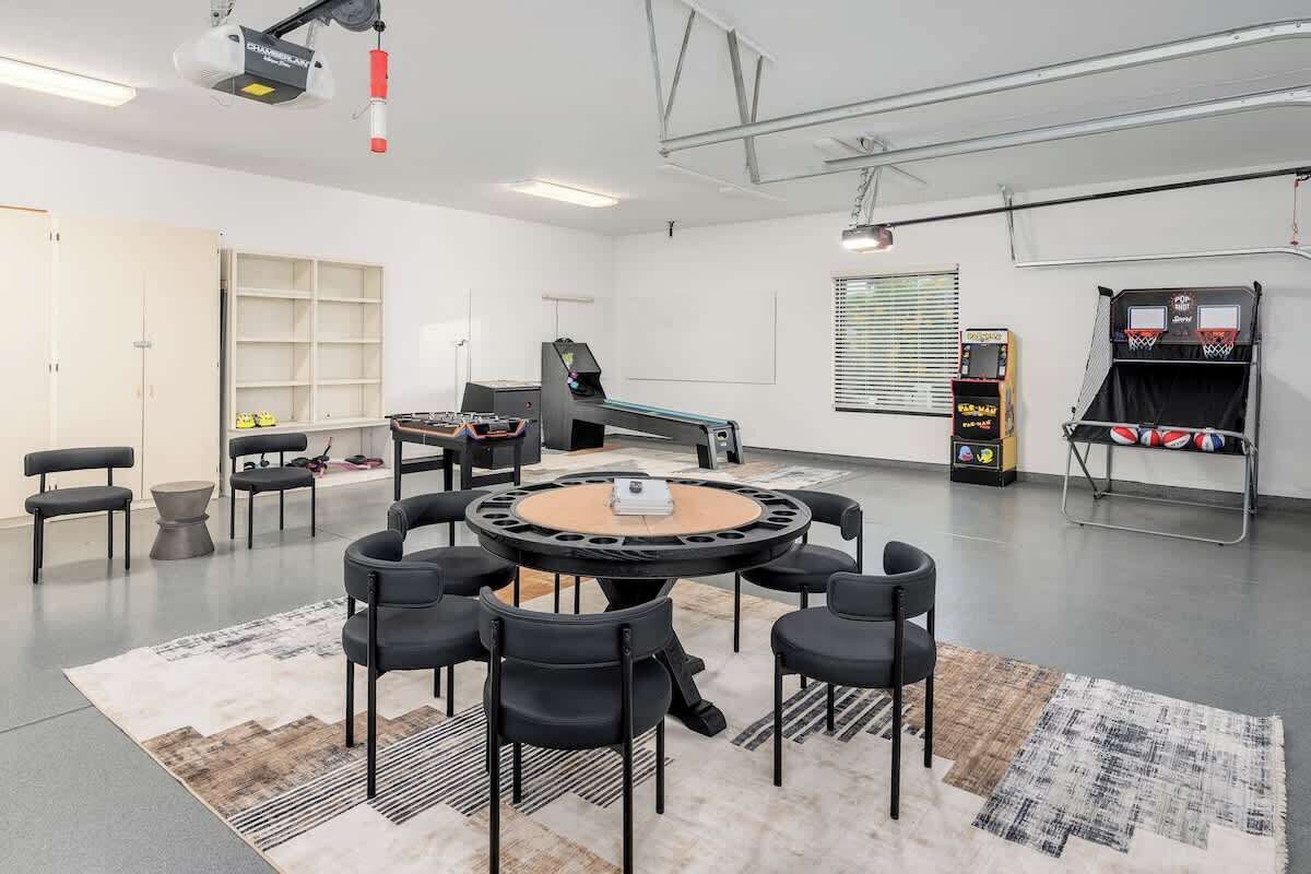A modern room with a round table, black chairs, a projector, a basketball arcade, and a window with blinds.