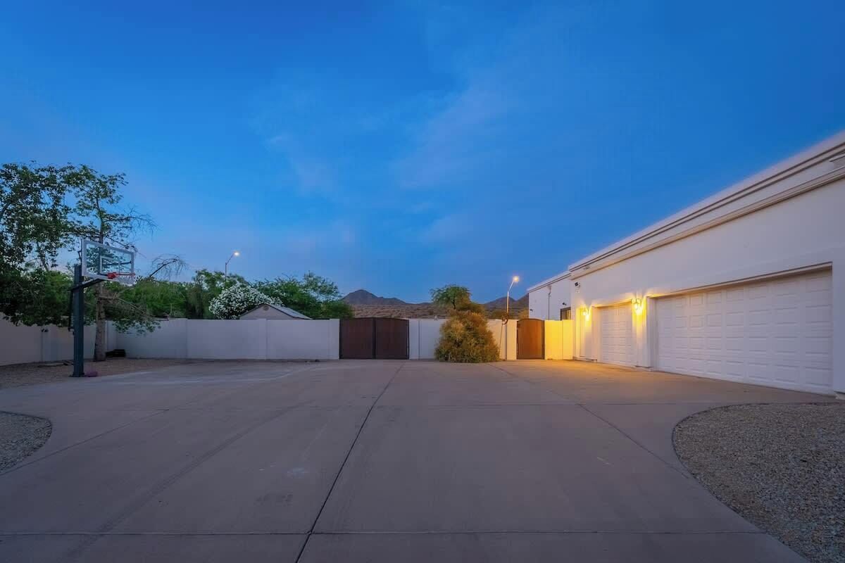 A residential driveway leading to a garage, surrounded by a white fence, with a basketball hoop on the left and a tree in the center.