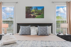 A bedroom with a bed, two bedside tables, a nightstand, a window with blinds, and a painting of a bridge. - 4BR Home Near MarshWalk + Pro Golf Simulator (Murrells Inlet)