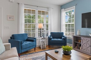 A living room with two blue armchairs, a small side table with a lamp, a coffee table with a plant, and a grey cabinet with books. - 4BR Home Near MarshWalk + Pro Golf Simulator (Murrells Inlet)