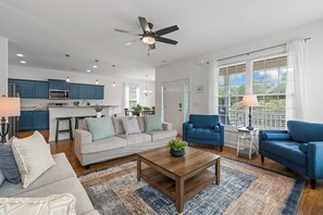 A modern living room with a sofa, coffee table, and blue armchairs. A kitchen with blue cabinets and a ceiling fan is visible in the background. - 4BR Home Near MarshWalk + Pro Golf Simulator (Murrells Inlet)