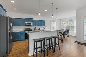 A modern kitchen with blue cabinets, a stainless steel refrigerator, a microwave, and a bar area with stools. - 4BR Home Near MarshWalk + Pro Golf Simulator (Murrells Inlet)