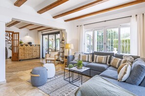 Un salon spacieux avec un canapé gris, des poutres en bois, une grande fenêtre et un meuble en bois.