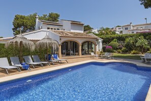Une piscine avec des chaises longues, un patio couvert et un bâtiment blanc doté d’un toit en tuiles.