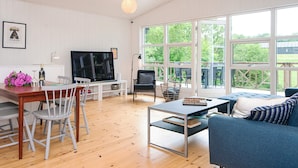 Un vaste espace de vie doté d’un plancher en bois, d’une table à manger avec des chaises, d’un canapé, d’un téléviseur et de grandes fenêtres offrant une vue sur la verdure.