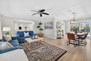 Ein geräumiges Wohnzimmer mit blauem Sofa, Couchtisch, Essbereich mit Tisch und Stühlen, Deckenventilator und großem Fenster mit Blick ins Freie.