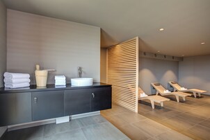Un baño moderno con tocador negro, lavabo blanco y toallas. Se observan separadores de madera en listones y una zona de estar con sillones reclinables.