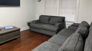 Ein Wohnzimmer mit einem grauen Ecksofa, einem kleinen Beistelltisch, einer Wanduhr und einem Fenster mit Jalousien.