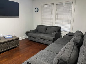 A living room with a grey sectional sofa, a small side table, a clock on the wall, and a window with blinds. - Vintage bungalow with quaint modern touches, minutes to Pensacola NAS front gate (pensacola)