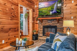 Ein gemütliches Blockhaus-Interieur mit Kamin, einer bequemen Couch, einem an der Wand montierten Fernseher und einem kleinen Tisch mit Kerzen.