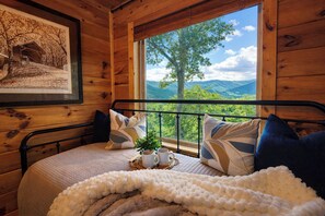 Ein Zimmer in einer Holzhütte mit einem Bett, Kissen, einem gerahmten Bild und einem Fenster mit Bergblick.