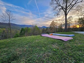 Un espace extérieur verdoyant, avec une couverture et des coussins, entouré d’arbres et offrant une vue sur des montagnes lointaines.