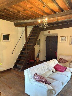 Een gezellige woonkamer met een houten trap, een witte bank met opvallende kussens en een kroonluchter.