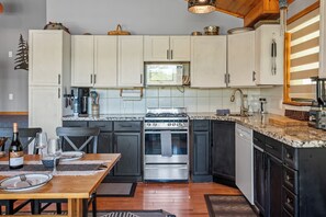 A kitchen with white cabinets, a stainless steel stove, and a microwave. There is a dining table with a bottle of wine and glasses.