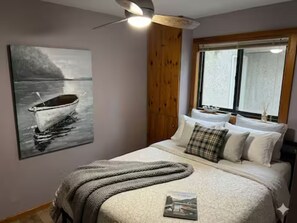 Une chambre à coucher avec un lit, des oreillers, une couverture, un tableau représentant un bateau, une fenêtre et un mur en bois.