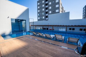 Piscina no terraço com espreguiçadeiras, uma parede de vidro e um edifício moderno ao fundo.