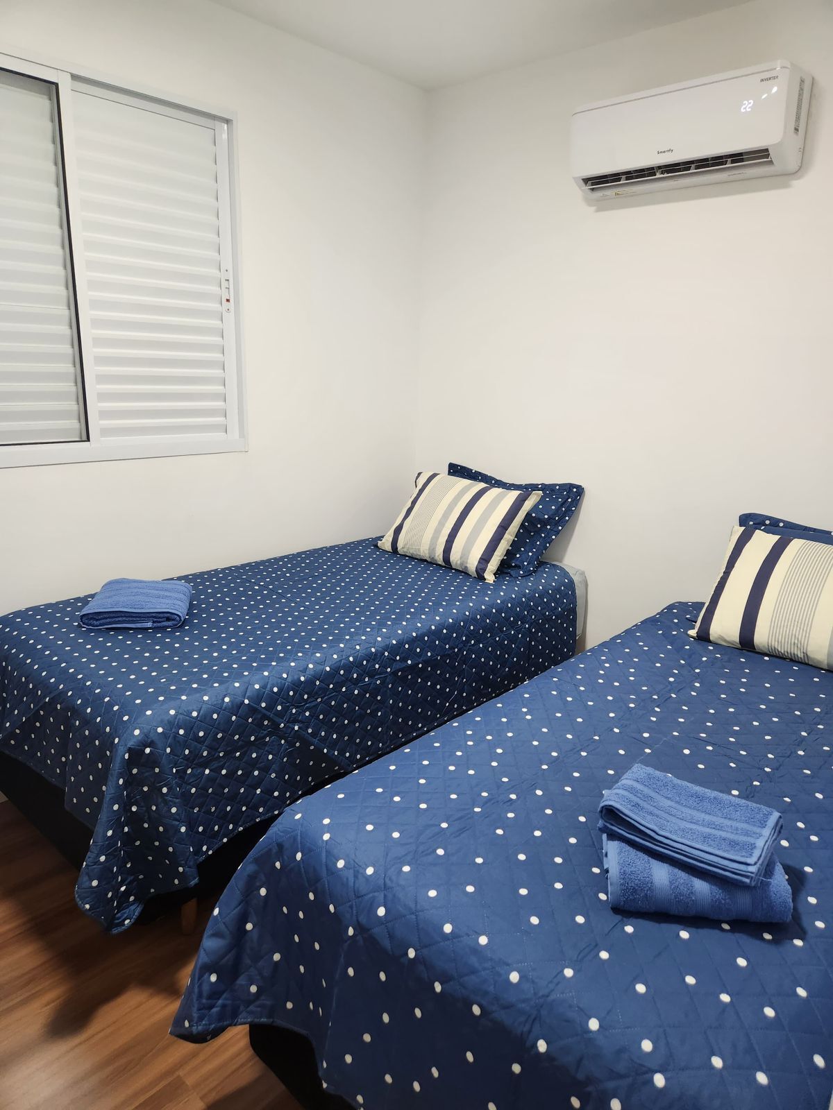 A room with two beds, each with a blue polka dot bedspread and matching pillows, a window with blinds, and a wall-mounted air conditioner.
