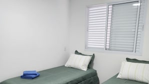 Ein Zimmer mit zwei Einzelbetten, jeweils mit einer grünen Tagesdecke und Kissen, einem Fenster mit Jalousien und einem Holzboden.