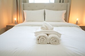 Een netjes opgemaakt bed met opgerolde handdoeken, een gestuufd hoofdbord en bedlampjes.