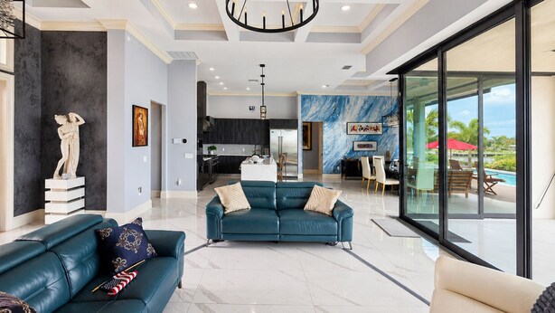 A spacious living area with a blue sofa, a marble floor, a modern kitchen, and a large glass door leading to a pool area.