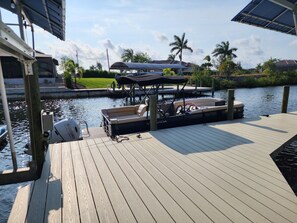 A dock with a boat, a covered seating area, and a building with a roof in the background.