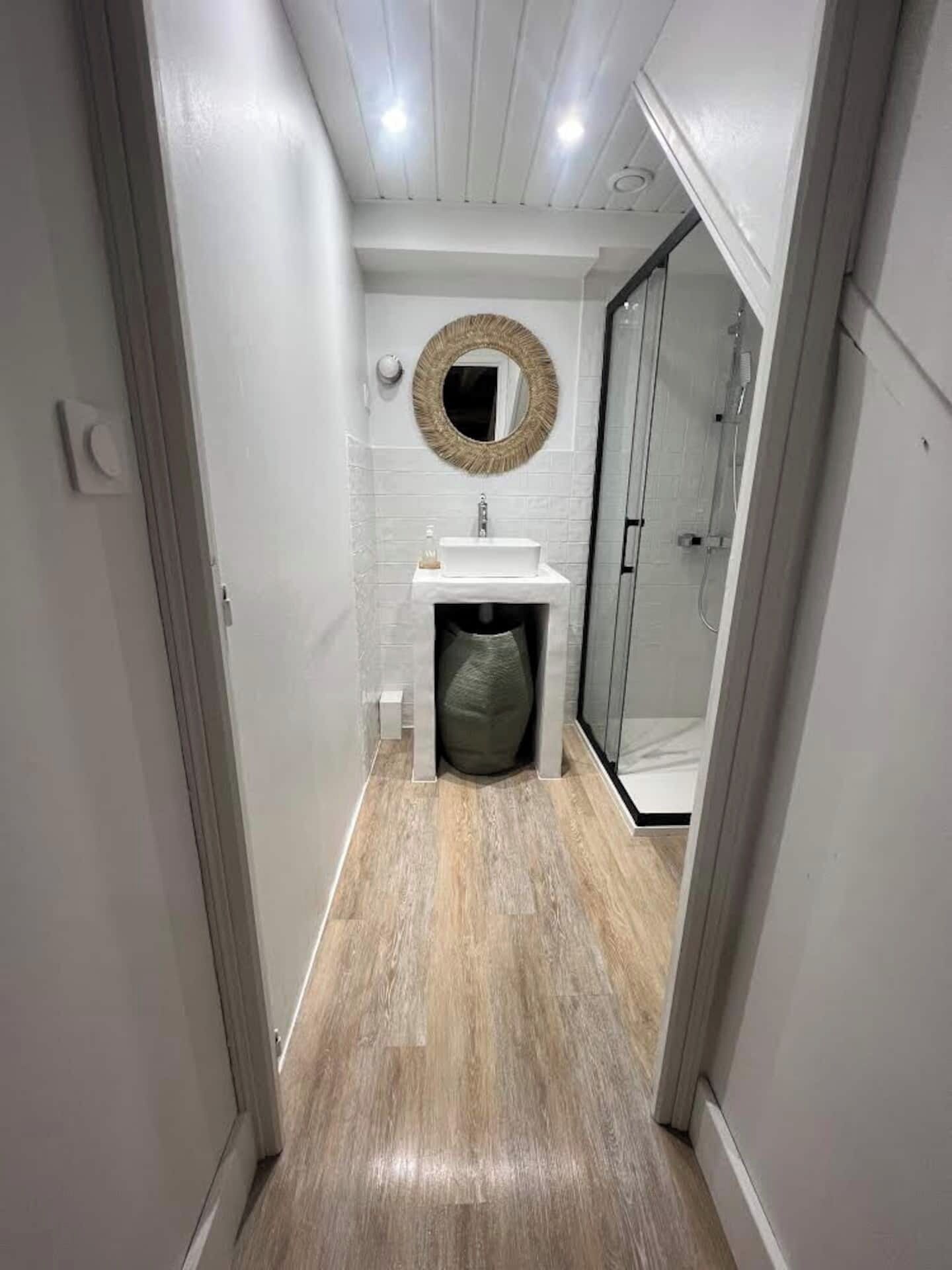 A narrow hallway leading to a bathroom with a white sink, a round mirror, and a shower area.