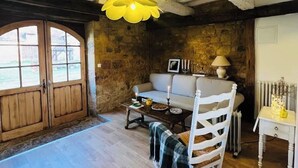 A rustic room with exposed beams, a stone wall, a wooden dining table, a white rocking chair, and a large window.